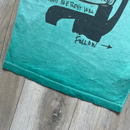 Dsquared2 SS2015 “Free Your Mind” T-shirt is a turquoise cotton tee with a washed, faded finish and tonal variation across the fabric. The front features a black Basquiat-inspired figure with “Free Your Mind and the Rest Will Follow” text in a contrasting design. The piece is made in Italy and includes darker tonal hems with a lighter center fade for a worn effect.