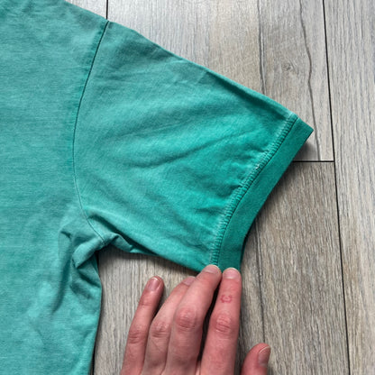 Dsquared2 SS2015 “Free Your Mind” T-shirt is a turquoise cotton tee with a washed, faded finish and tonal variation across the fabric. The front features a black Basquiat-inspired figure with “Free Your Mind and the Rest Will Follow” text in a contrasting design. The piece is made in Italy and includes darker tonal hems with a lighter center fade for a worn effect.