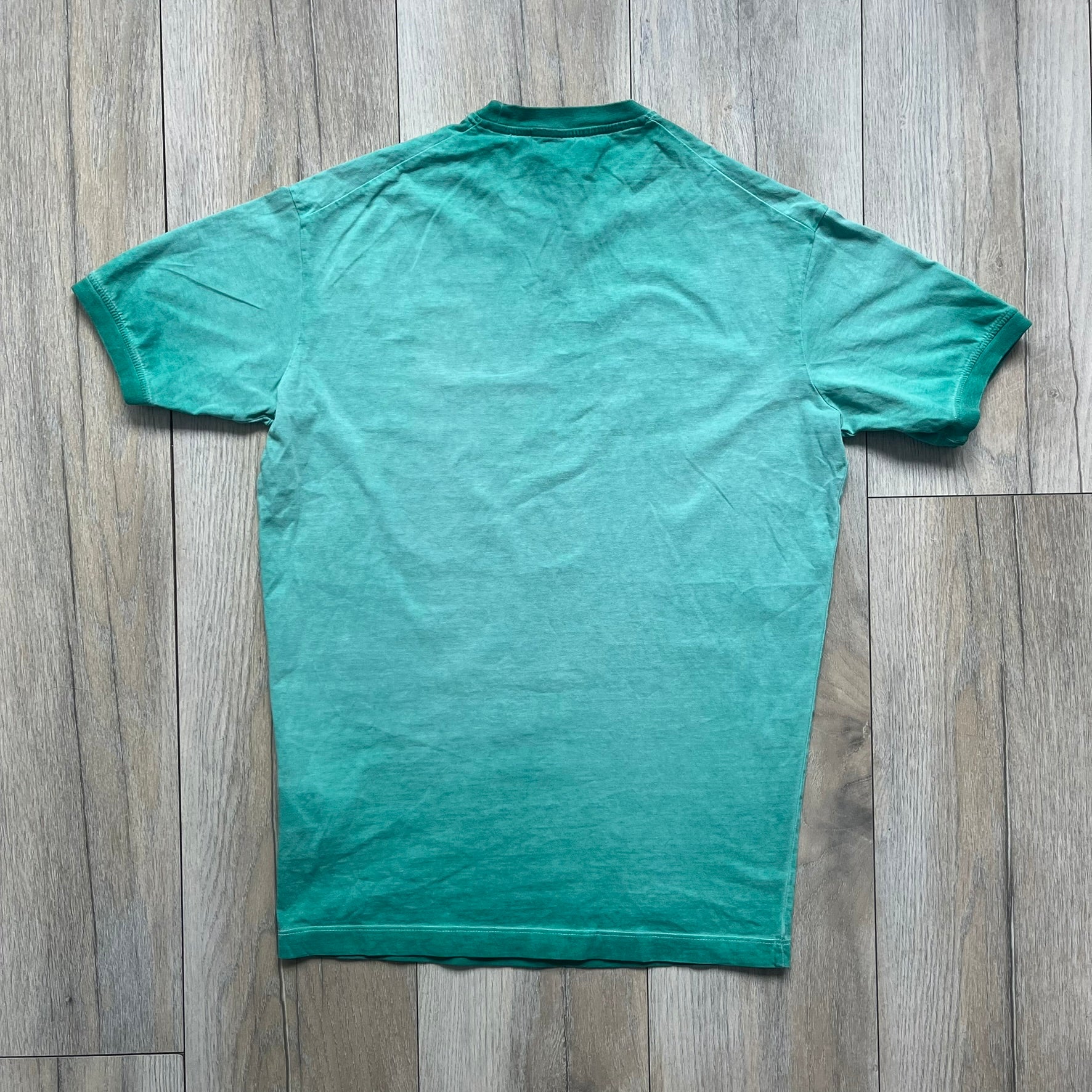 Dsquared2 SS2015 “Free Your Mind” T-shirt is a turquoise cotton tee with a washed, faded finish and tonal variation across the fabric. The front features a black Basquiat-inspired figure with “Free Your Mind and the Rest Will Follow” text in a contrasting design. The piece is made in Italy and includes darker tonal hems with a lighter center fade for a worn effect.