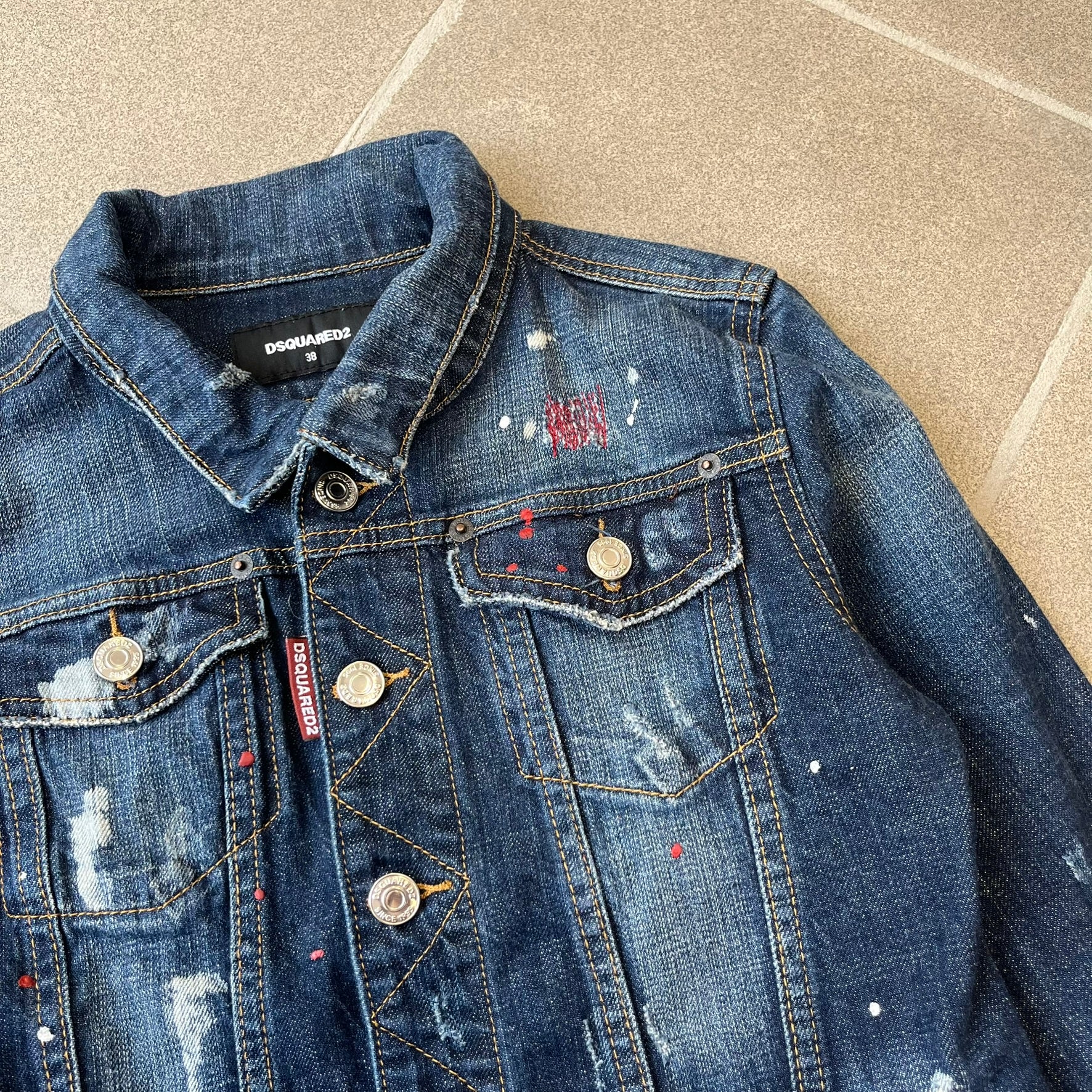 Dsquared2 SS15 Rebel Monkey Denim Jacket features a denim construction with two chest pockets on the front. The back has a large Rebel Monkey graphic in white, blue, and red, with black Dsquared2 text above it. The jacket includes intentional tears, paint splatters, and other distressed details. Made in Italy as part of the Dsquared2 collaboration with Basquiat.

