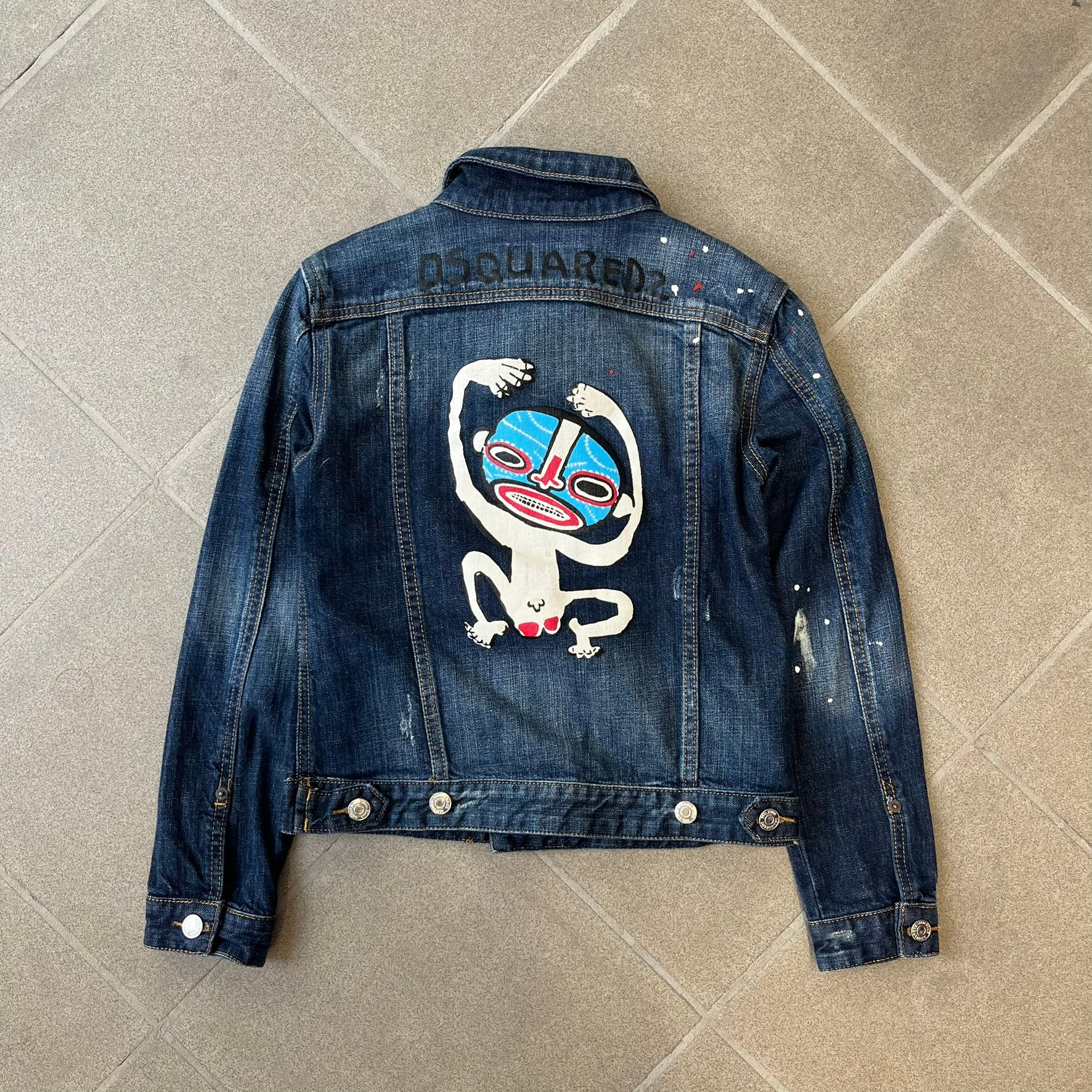 Dsquared2 SS15 Rebel Monkey Denim Jacket features a denim construction with two chest pockets on the front. The back has a large Rebel Monkey graphic in white, blue, and red, with black Dsquared2 text above it. The jacket includes intentional tears, paint splatters, and other distressed details. Made in Italy as part of the Dsquared2 collaboration with Basquiat.
