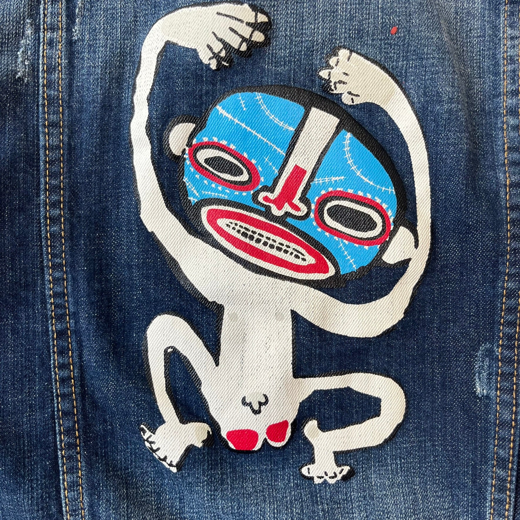 Dsquared2 SS15 Rebel Monkey Denim Jacket features a denim construction with two chest pockets on the front. The back has a large Rebel Monkey graphic in white, blue, and red, with black Dsquared2 text above it. The jacket includes intentional tears, paint splatters, and other distressed details. Made in Italy as part of the Dsquared2 collaboration with Basquiat.

