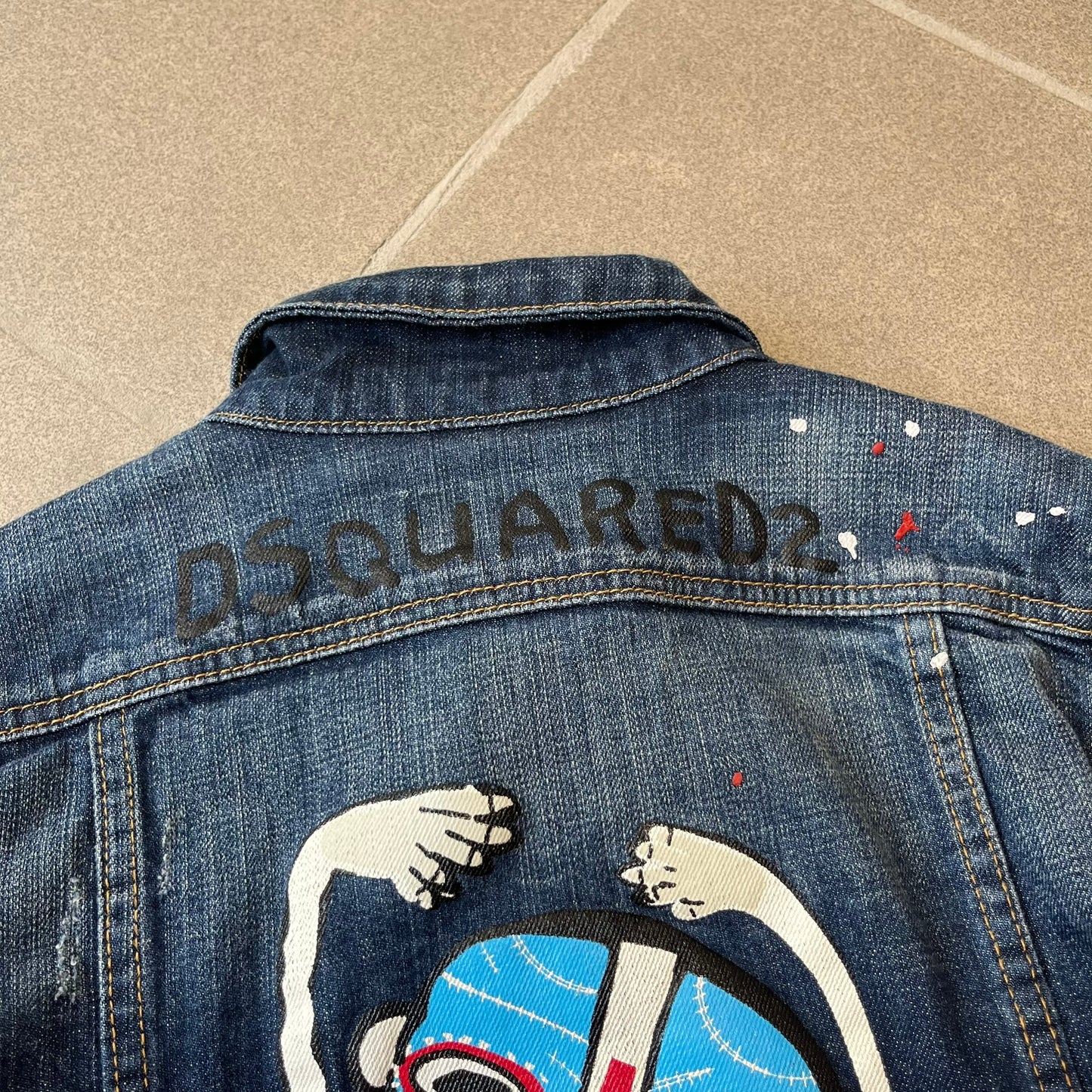 Dsquared2 SS15 Rebel Monkey Denim Jacket features a denim construction with two chest pockets on the front. The back has a large Rebel Monkey graphic in white, blue, and red, with black Dsquared2 text above it. The jacket includes intentional tears, paint splatters, and other distressed details. Made in Italy as part of the Dsquared2 collaboration with Basquiat.
