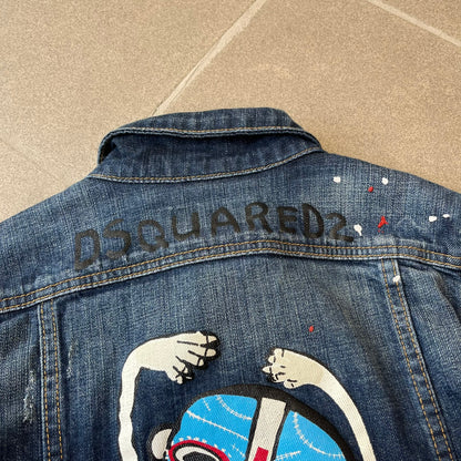 Dsquared2 SS15 Rebel Monkey Denim Jacket features a denim construction with two chest pockets on the front. The back has a large Rebel Monkey graphic in white, blue, and red, with black Dsquared2 text above it. The jacket includes intentional tears, paint splatters, and other distressed details. Made in Italy as part of the Dsquared2 collaboration with Basquiat.
