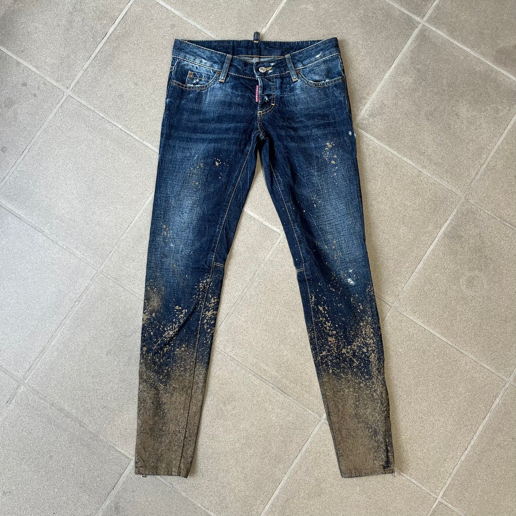 Dsquared2 SS2012 Mud Splash Jeans are runway bottoms from the Spring/Summer 2012 collection, made in Italy from 100% cotton. The jeans feature a deliberate two-tone design with a clean blue-denim upper section transitioning into a heavy brown “mud” effect on the lower half. They include branded buttons and rivets, a Dsquared2 back patch, and a small red maple leaf metal pin on the back pocket.