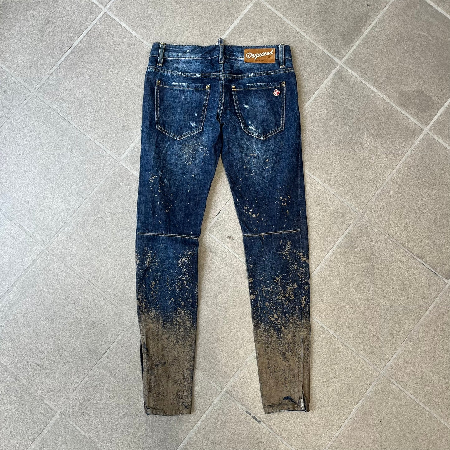 Dsquared2 SS2012 Mud Splash Jeans are runway bottoms from the Spring/Summer 2012 collection, made in Italy from 100% cotton. The jeans feature a deliberate two-tone design with a clean blue-denim upper section transitioning into a heavy brown “mud” effect on the lower half. They include branded buttons and rivets, a Dsquared2 back patch, and a small red maple leaf metal pin on the back pocket.