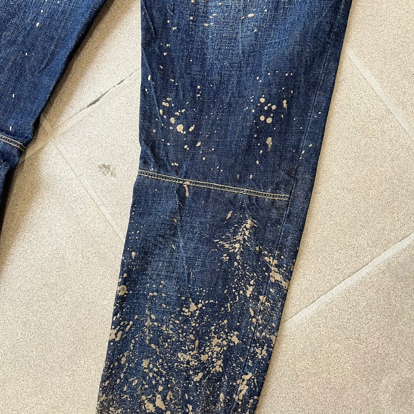 Dsquared2 SS2012 Mud Splash Jeans are runway bottoms from the Spring/Summer 2012 collection, made in Italy from 100% cotton. The jeans feature a deliberate two-tone design with a clean blue-denim upper section transitioning into a heavy brown “mud” effect on the lower half. They include branded buttons and rivets, a Dsquared2 back patch, and a small red maple leaf metal pin on the back pocket.