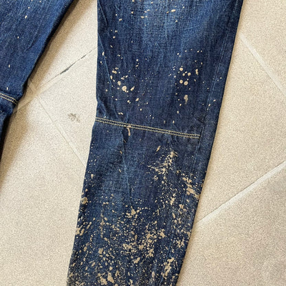 Dsquared2 SS2012 Mud Splash Jeans are runway bottoms from the Spring/Summer 2012 collection, made in Italy from 100% cotton. The jeans feature a deliberate two-tone design with a clean blue-denim upper section transitioning into a heavy brown “mud” effect on the lower half. They include branded buttons and rivets, a Dsquared2 back patch, and a small red maple leaf metal pin on the back pocket.