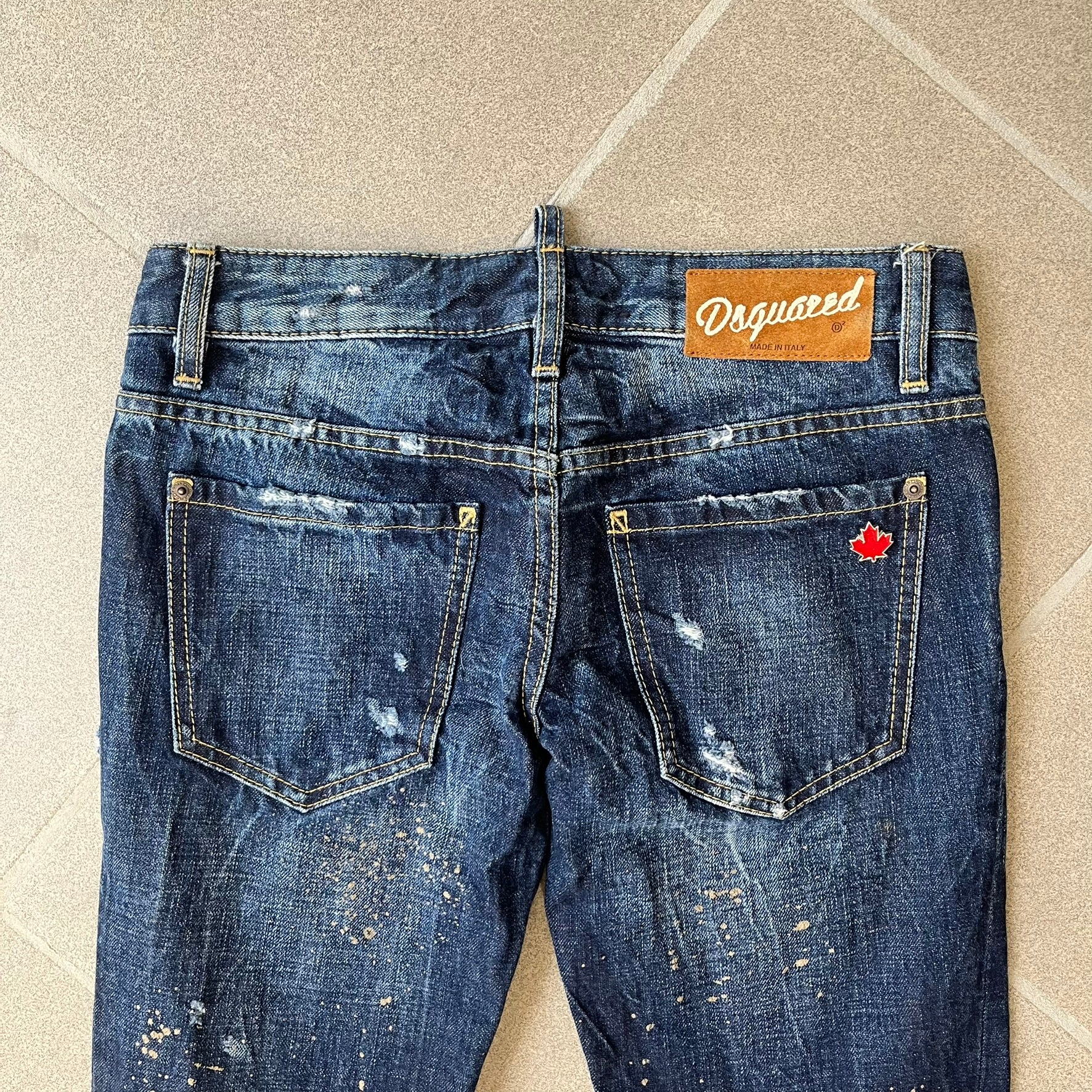 Dsquared2 SS2012 Mud Splash Jeans are runway bottoms from the Spring/Summer 2012 collection, made in Italy from 100% cotton. The jeans feature a deliberate two-tone design with a clean blue-denim upper section transitioning into a heavy brown “mud” effect on the lower half. They include branded buttons and rivets, a Dsquared2 back patch, and a small red maple leaf metal pin on the back pocket.