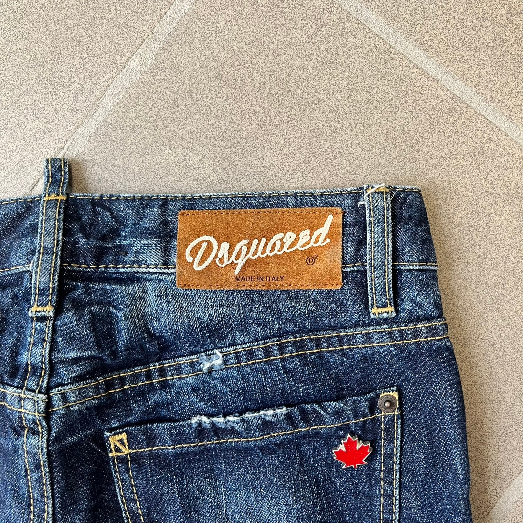 Dsquared2 SS2012 Mud Splash Jeans are runway bottoms from the Spring/Summer 2012 collection, made in Italy from 100% cotton. The jeans feature a deliberate two-tone design with a clean blue-denim upper section transitioning into a heavy brown “mud” effect on the lower half. They include branded buttons and rivets, a Dsquared2 back patch, and a small red maple leaf metal pin on the back pocket.