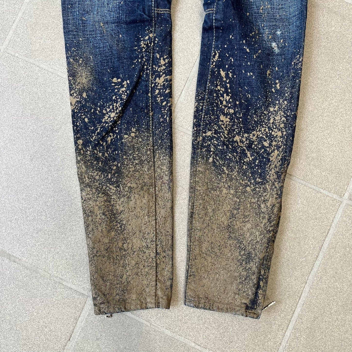 Dsquared2 SS2012 Mud Splash Jeans are runway bottoms from the Spring/Summer 2012 collection, made in Italy from 100% cotton. The jeans feature a deliberate two-tone design with a clean blue-denim upper section transitioning into a heavy brown “mud” effect on the lower half. They include branded buttons and rivets, a Dsquared2 back patch, and a small red maple leaf metal pin on the back pocket.