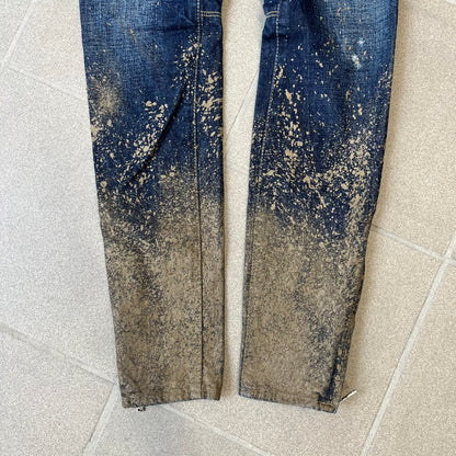 Dsquared2 SS2012 Mud Splash Jeans are runway bottoms from the Spring/Summer 2012 collection, made in Italy from 100% cotton. The jeans feature a deliberate two-tone design with a clean blue-denim upper section transitioning into a heavy brown “mud” effect on the lower half. They include branded buttons and rivets, a Dsquared2 back patch, and a small red maple leaf metal pin on the back pocket.