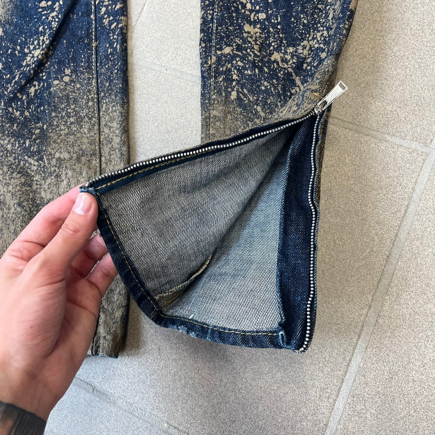 Dsquared2 SS2012 Mud Splash Jeans are runway bottoms from the Spring/Summer 2012 collection, made in Italy from 100% cotton. The jeans feature a deliberate two-tone design with a clean blue-denim upper section transitioning into a heavy brown “mud” effect on the lower half. They include branded buttons and rivets, a Dsquared2 back patch, and a small red maple leaf metal pin on the back pocket.