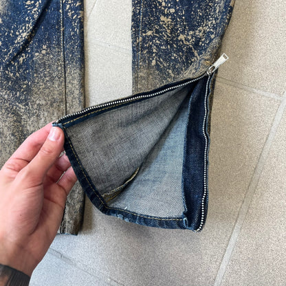 Dsquared2 SS2012 Mud Splash Jeans are runway bottoms from the Spring/Summer 2012 collection, made in Italy from 100% cotton. The jeans feature a deliberate two-tone design with a clean blue-denim upper section transitioning into a heavy brown “mud” effect on the lower half. They include branded buttons and rivets, a Dsquared2 back patch, and a small red maple leaf metal pin on the back pocket.