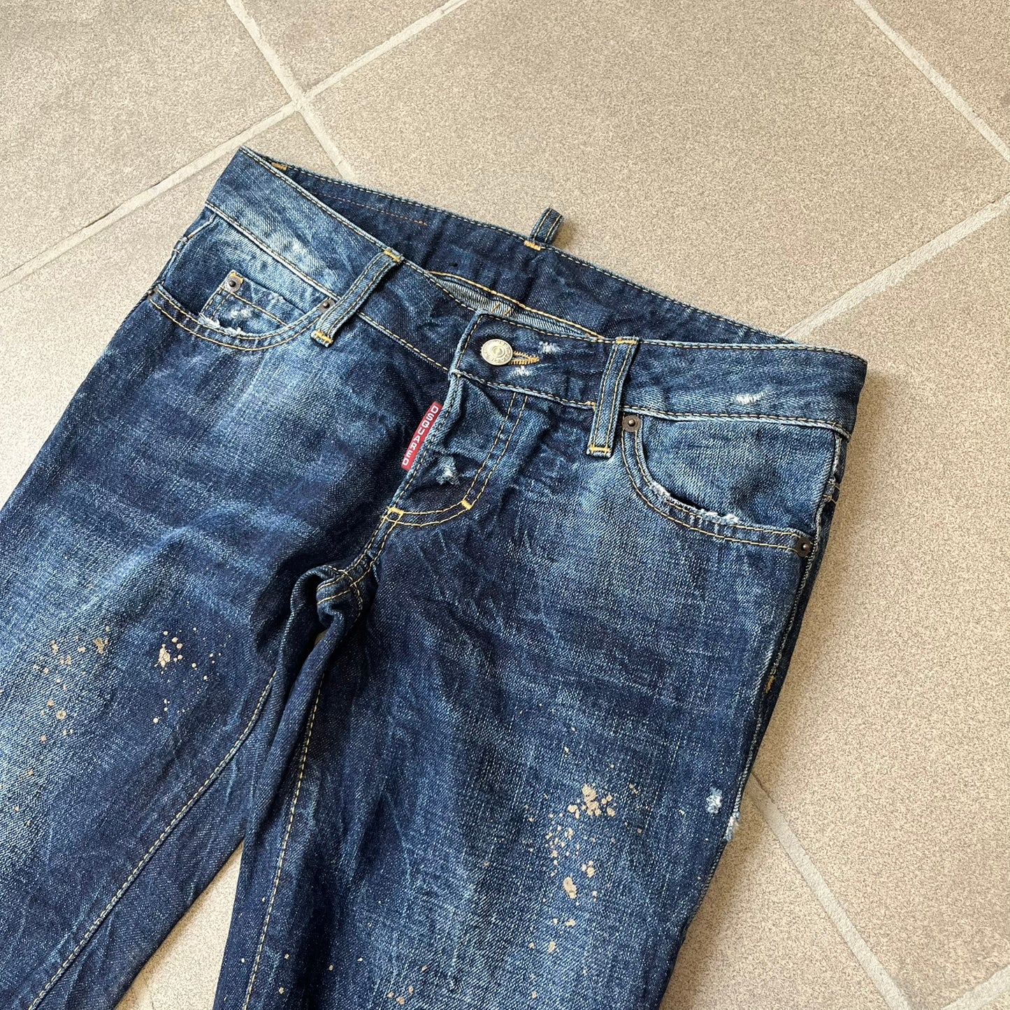 Dsquared2 SS2012 Mud Splash Jeans are runway bottoms from the Spring/Summer 2012 collection, made in Italy from 100% cotton. The jeans feature a deliberate two-tone design with a clean blue-denim upper section transitioning into a heavy brown “mud” effect on the lower half. They include branded buttons and rivets, a Dsquared2 back patch, and a small red maple leaf metal pin on the back pocket.