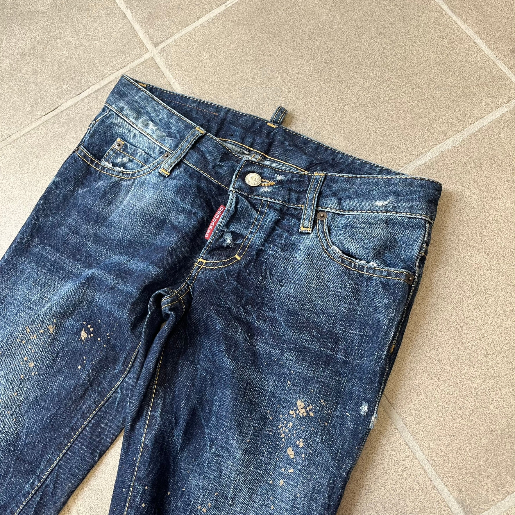 Dsquared2 SS2012 Mud Splash Jeans are runway bottoms from the Spring/Summer 2012 collection, made in Italy from 100% cotton. The jeans feature a deliberate two-tone design with a clean blue-denim upper section transitioning into a heavy brown “mud” effect on the lower half. They include branded buttons and rivets, a Dsquared2 back patch, and a small red maple leaf metal pin on the back pocket.