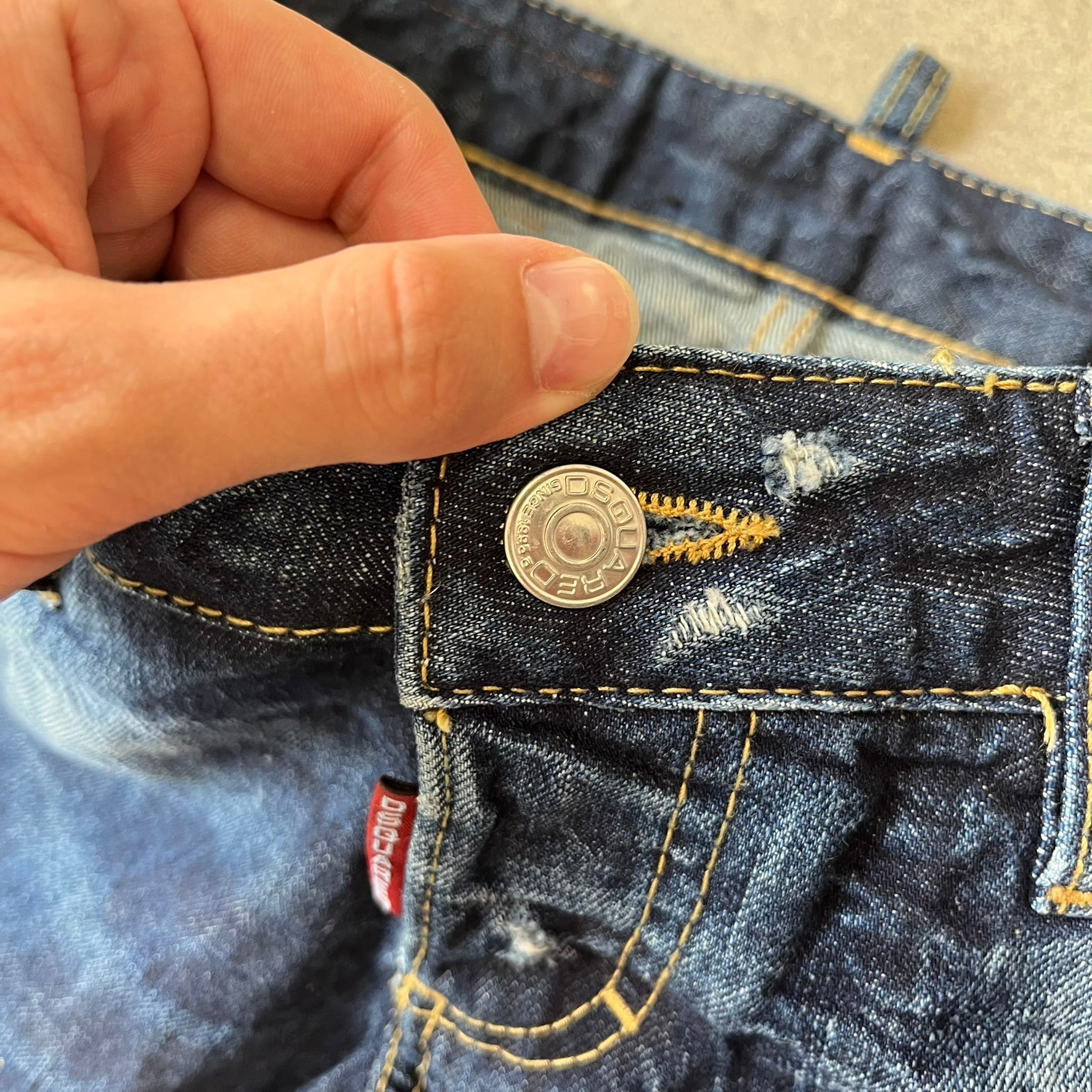 Dsquared2 SS2012 Mud Splash Jeans are runway bottoms from the Spring/Summer 2012 collection, made in Italy from 100% cotton. The jeans feature a deliberate two-tone design with a clean blue-denim upper section transitioning into a heavy brown “mud” effect on the lower half. They include branded buttons and rivets, a Dsquared2 back patch, and a small red maple leaf metal pin on the back pocket.