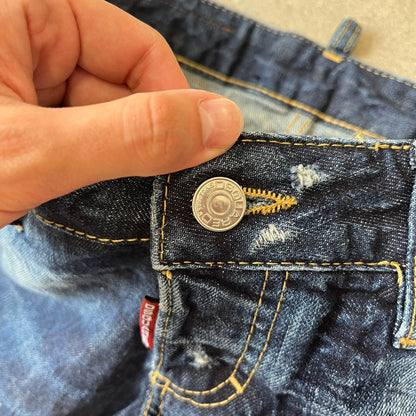 Dsquared2 SS2012 Mud Splash Jeans are runway bottoms from the Spring/Summer 2012 collection, made in Italy from 100% cotton. The jeans feature a deliberate two-tone design with a clean blue-denim upper section transitioning into a heavy brown “mud” effect on the lower half. They include branded buttons and rivets, a Dsquared2 back patch, and a small red maple leaf metal pin on the back pocket.