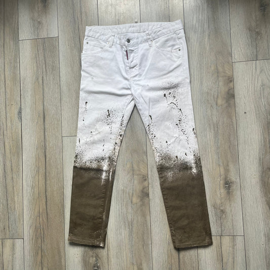 Dsquared2 SS2012 White Mud Jeans are runway pieces from the Spring/Summer 2012 collection, made in Italy from 98% cotton and 2% elastane. The jeans feature a deliberate two-tone design with a clean white upper section transitioning into a heavy brown “mud” effect on the lower half. They include branded buttons and rivets, a Dsquared2 front patch, and a small red maple leaf metal pin on the back pocket. The leg opening measures approximately 17 cm, with a slightly stretchy slim fit construction.