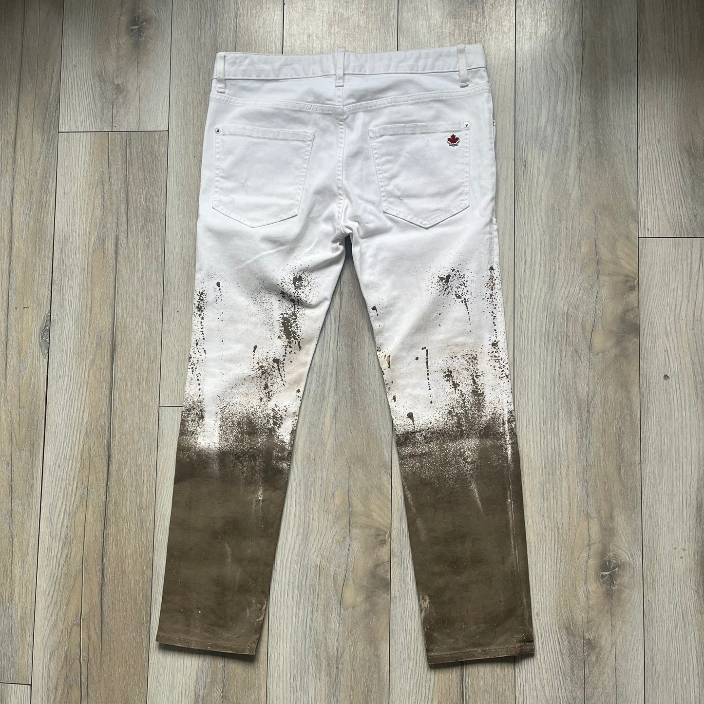 Dsquared2 SS2012 White Mud Jeans are runway pieces from the Spring/Summer 2012 collection, made in Italy from 98% cotton and 2% elastane. The jeans feature a deliberate two-tone design with a clean white upper section transitioning into a heavy brown “mud” effect on the lower half. They include branded buttons and rivets, a Dsquared2 front patch, and a small red maple leaf metal pin on the back pocket. The leg opening measures approximately 17 cm, with a slightly stretchy slim fit construction.