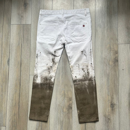 Dsquared2 SS2012 White Mud Jeans are runway pieces from the Spring/Summer 2012 collection, made in Italy from 98% cotton and 2% elastane. The jeans feature a deliberate two-tone design with a clean white upper section transitioning into a heavy brown “mud” effect on the lower half. They include branded buttons and rivets, a Dsquared2 front patch, and a small red maple leaf metal pin on the back pocket. The leg opening measures approximately 17 cm, with a slightly stretchy slim fit construction.