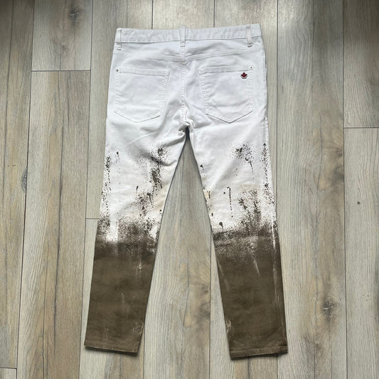 Dsquared2 SS2012 White Mud Jeans are runway pieces from the Spring/Summer 2012 collection, made in Italy from 98% cotton and 2% elastane. The jeans feature a deliberate two-tone design with a clean white upper section transitioning into a heavy brown “mud” effect on the lower half. They include branded buttons and rivets, a Dsquared2 front patch, and a small red maple leaf metal pin on the back pocket. The leg opening measures approximately 17 cm, with a slightly stretchy slim fit construction.