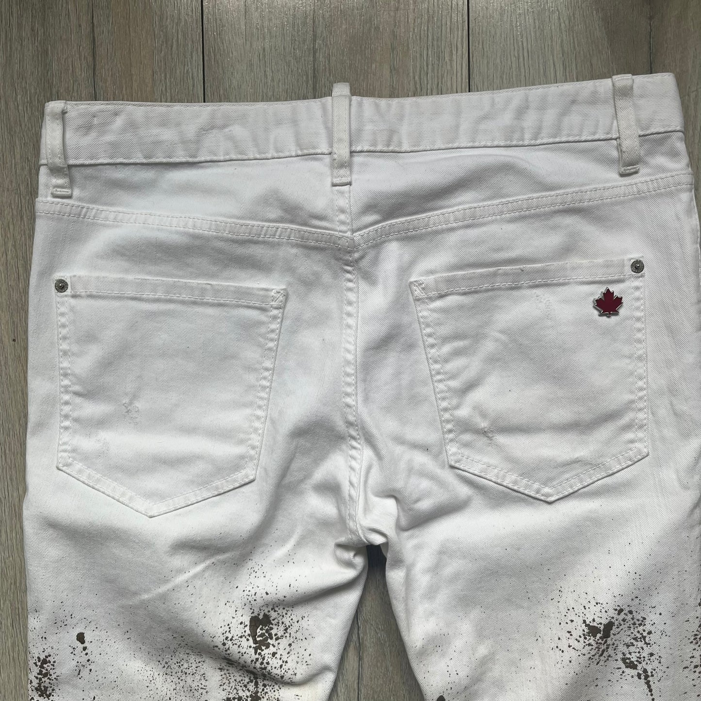Dsquared2 SS2012 White Mud Jeans are runway pieces from the Spring/Summer 2012 collection, made in Italy from 98% cotton and 2% elastane. The jeans feature a deliberate two-tone design with a clean white upper section transitioning into a heavy brown “mud” effect on the lower half. They include branded buttons and rivets, a Dsquared2 front patch, and a small red maple leaf metal pin on the back pocket. The leg opening measures approximately 17 cm, with a slightly stretchy slim fit construction.