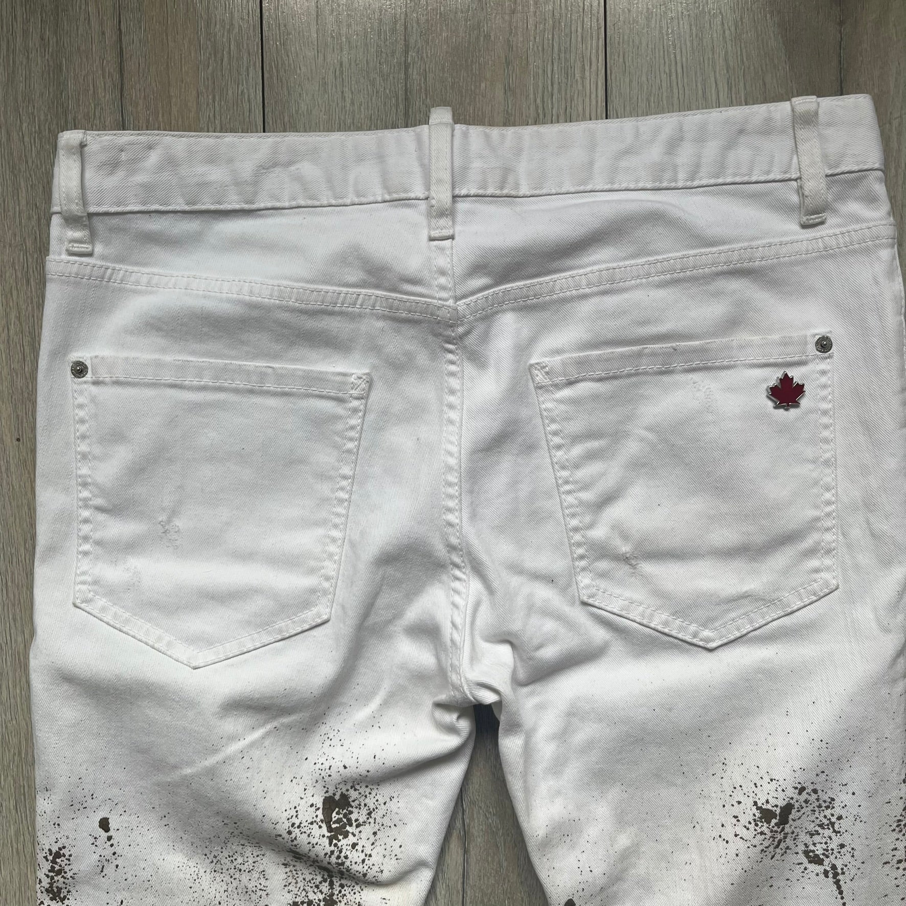 Dsquared2 SS2012 White Mud Jeans are runway pieces from the Spring/Summer 2012 collection, made in Italy from 98% cotton and 2% elastane. The jeans feature a deliberate two-tone design with a clean white upper section transitioning into a heavy brown “mud” effect on the lower half. They include branded buttons and rivets, a Dsquared2 front patch, and a small red maple leaf metal pin on the back pocket. The leg opening measures approximately 17 cm, with a slightly stretchy slim fit construction.