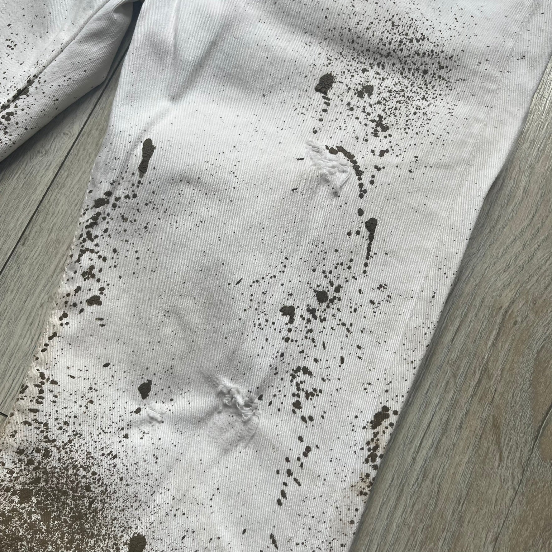 Dsquared2 SS2012 White Mud Jeans are runway pieces from the Spring/Summer 2012 collection, made in Italy from 98% cotton and 2% elastane. The jeans feature a deliberate two-tone design with a clean white upper section transitioning into a heavy brown “mud” effect on the lower half. They include branded buttons and rivets, a Dsquared2 front patch, and a small red maple leaf metal pin on the back pocket. The leg opening measures approximately 17 cm, with a slightly stretchy slim fit construction.