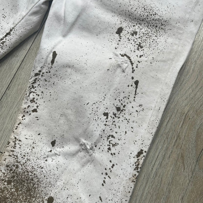 Dsquared2 SS2012 White Mud Jeans are runway pieces from the Spring/Summer 2012 collection, made in Italy from 98% cotton and 2% elastane. The jeans feature a deliberate two-tone design with a clean white upper section transitioning into a heavy brown “mud” effect on the lower half. They include branded buttons and rivets, a Dsquared2 front patch, and a small red maple leaf metal pin on the back pocket. The leg opening measures approximately 17 cm, with a slightly stretchy slim fit construction.