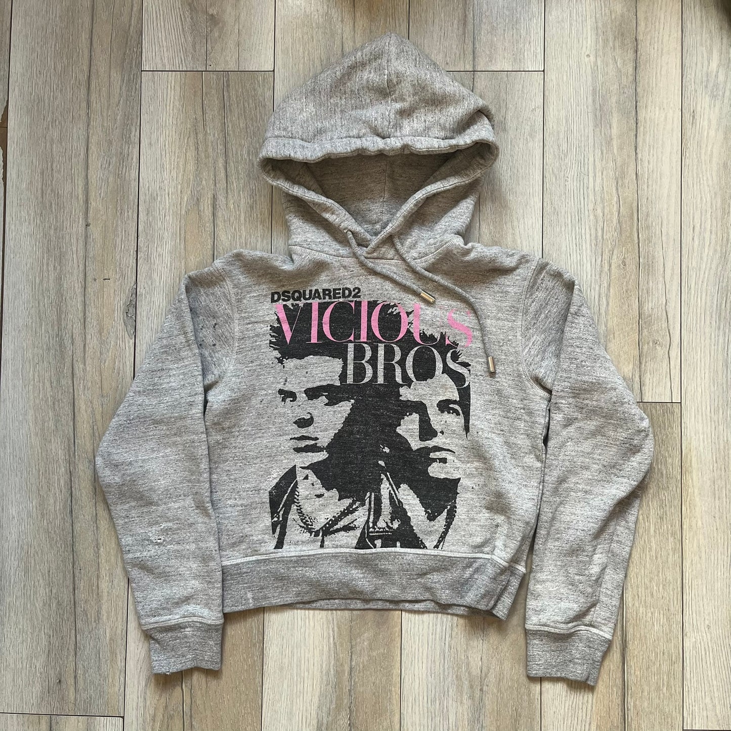 Dsquared2 Vicious Bros SS2019 Hoodie is a grey cotton hoodie made in Italy. It features a hood with drawstring fastening, long sleeves, and ribbed cuffs and hem. The front includes a printed graphic of two outlined figures with a small Dsquared2 logo and pink “Vicious Bros” text above them. The hoodie has intentional distressing throughout the fabric.
