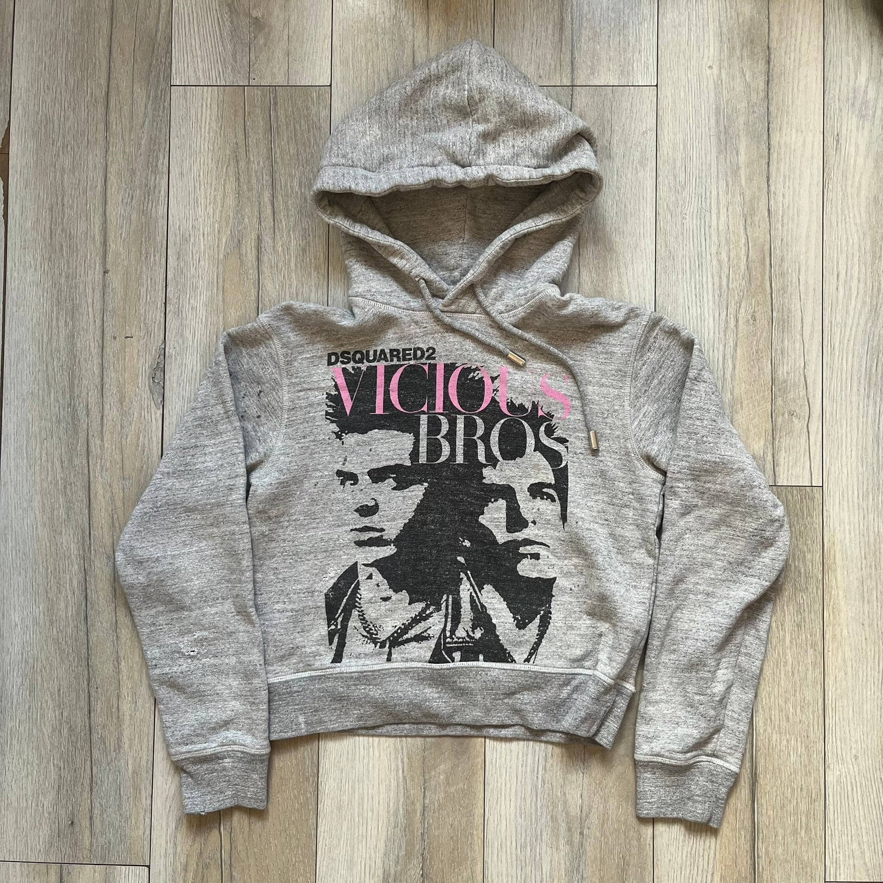 Dsquared2 Vicious Bros SS2019 Hoodie is a grey cotton hoodie made in Italy. It features a hood with drawstring fastening, long sleeves, and ribbed cuffs and hem. The front includes a printed graphic of two outlined figures with a small Dsquared2 logo and pink “Vicious Bros” text above them. The hoodie has intentional distressing throughout the fabric.
