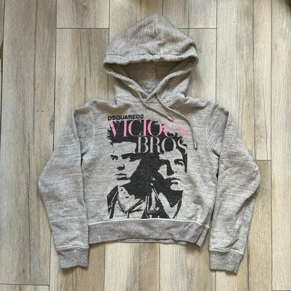 Dsquared2 Vicious Bros SS2019 Hoodie is a grey cotton hoodie made in Italy. It features a hood with drawstring fastening, long sleeves, and ribbed cuffs and hem. The front includes a printed graphic of two outlined figures with a small Dsquared2 logo and pink “Vicious Bros” text above them. The hoodie has intentional distressing throughout the fabric.
