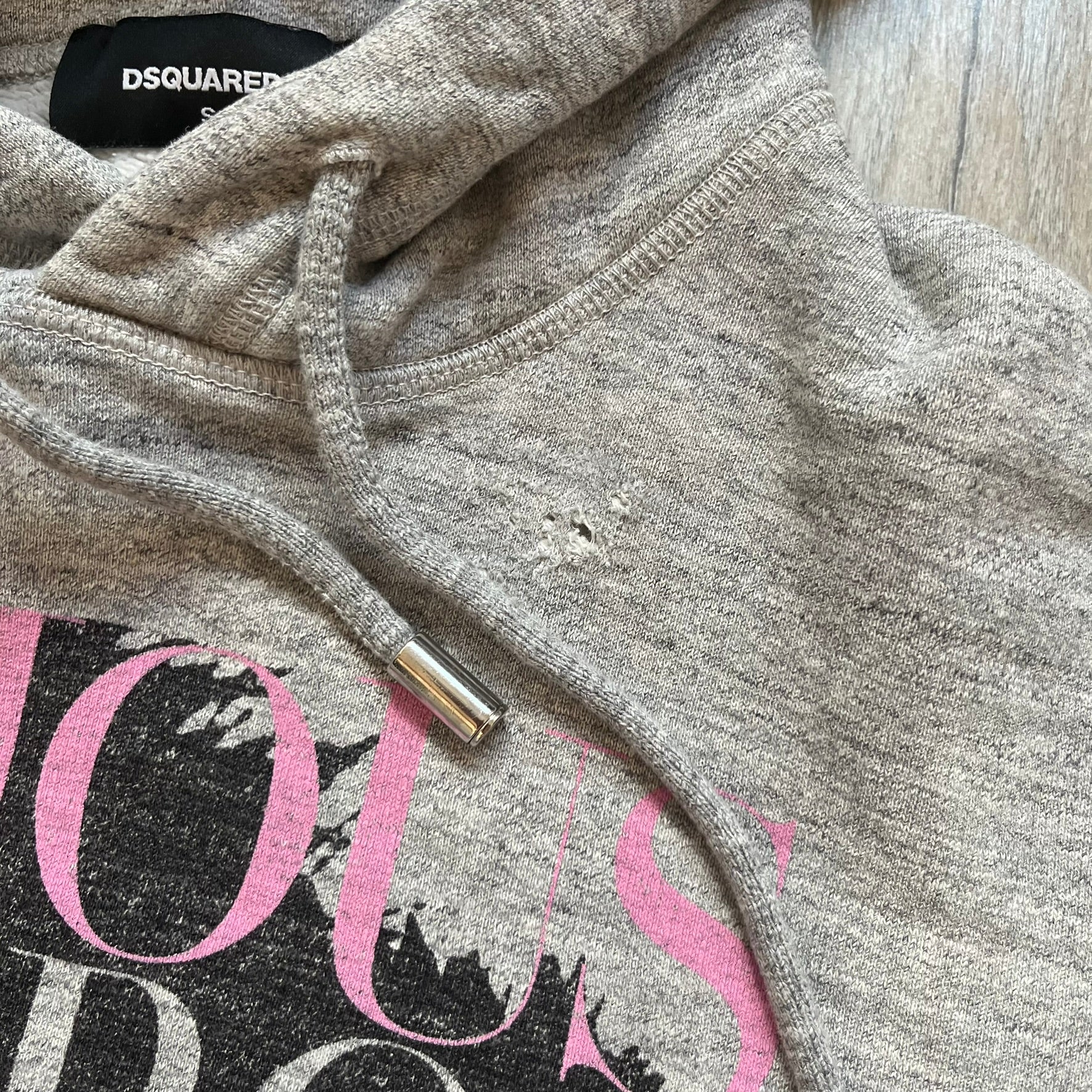 Dsquared2 Vicious Bros SS2019 Hoodie is a grey cotton hoodie made in Italy. It features a hood with drawstring fastening, long sleeves, and ribbed cuffs and hem. The front includes a printed graphic of two outlined figures with a small Dsquared2 logo and pink “Vicious Bros” text above them. The hoodie has intentional distressing throughout the fabric.
