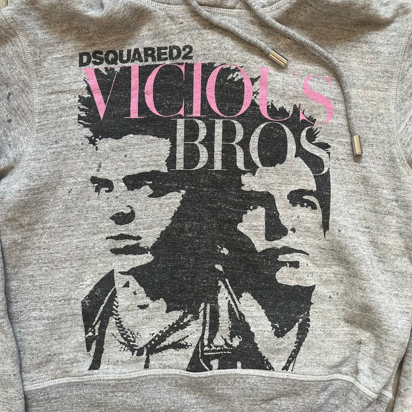 Dsquared2 Vicious Bros SS2019 Hoodie is a grey cotton hoodie made in Italy. It features a hood with drawstring fastening, long sleeves, and ribbed cuffs and hem. The front includes a printed graphic of two outlined figures with a small Dsquared2 logo and pink “Vicious Bros” text above them. The hoodie has intentional distressing throughout the fabric.
