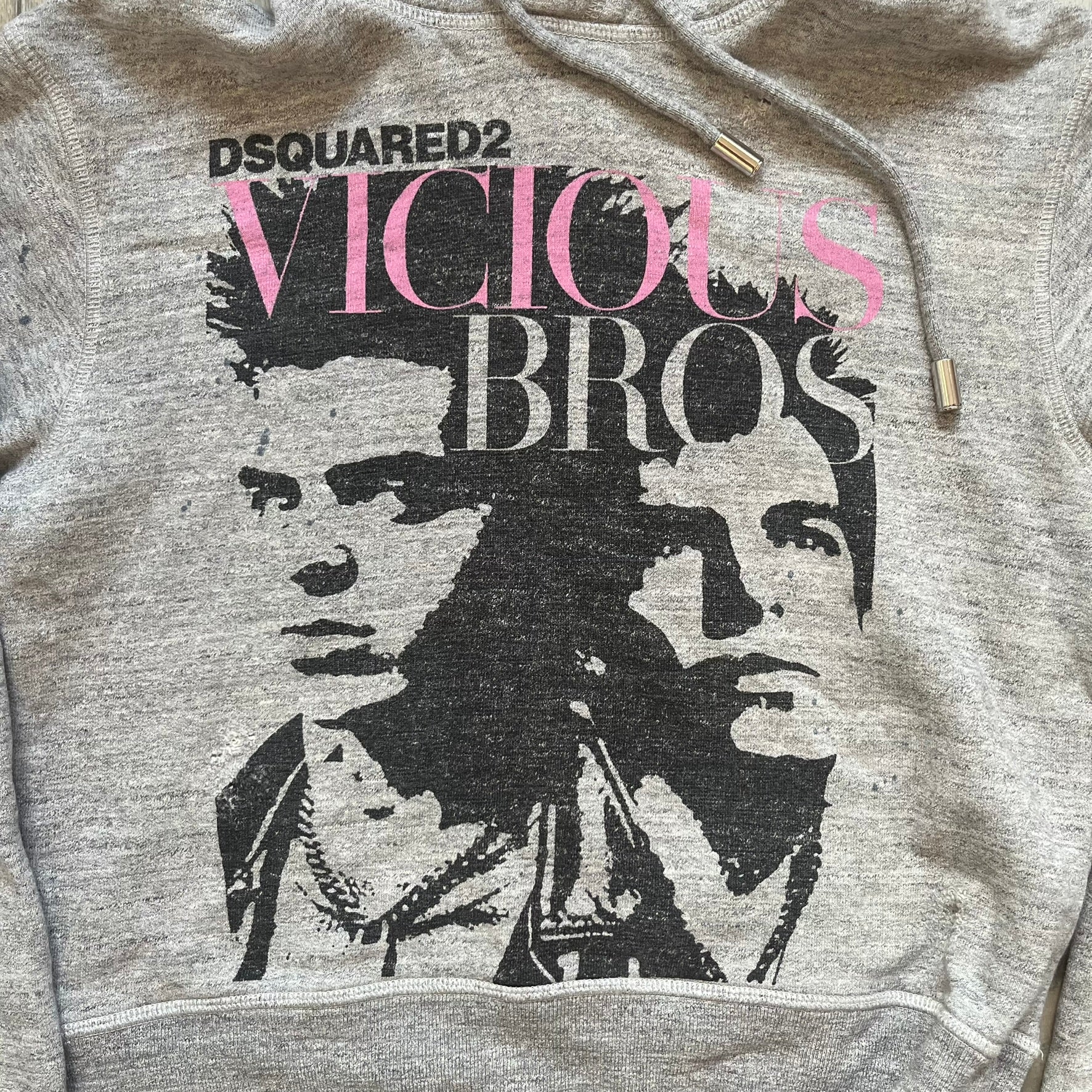 Dsquared2 Vicious Bros SS2019 Hoodie is a grey cotton hoodie made in Italy. It features a hood with drawstring fastening, long sleeves, and ribbed cuffs and hem. The front includes a printed graphic of two outlined figures with a small Dsquared2 logo and pink “Vicious Bros” text above them. The hoodie has intentional distressing throughout the fabric.
