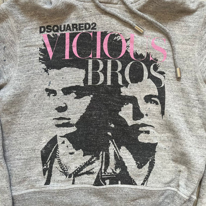 Dsquared2 Vicious Bros SS2019 Hoodie is a grey cotton hoodie made in Italy. It features a hood with drawstring fastening, long sleeves, and ribbed cuffs and hem. The front includes a printed graphic of two outlined figures with a small Dsquared2 logo and pink “Vicious Bros” text above them. The hoodie has intentional distressing throughout the fabric.
