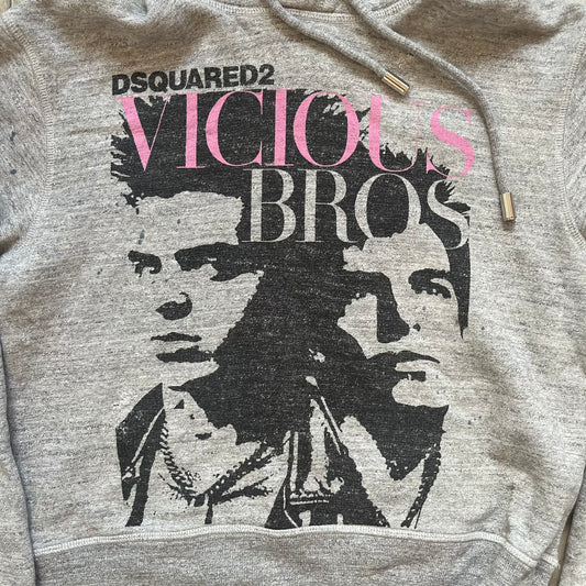 Dsquared2 Vicious Bros SS2019 Hoodie is a grey cotton hoodie made in Italy. It features a hood with drawstring fastening, long sleeves, and ribbed cuffs and hem. The front includes a printed graphic of two outlined figures with a small Dsquared2 logo and pink “Vicious Bros” text above them. The hoodie has intentional distressing throughout the fabric.
