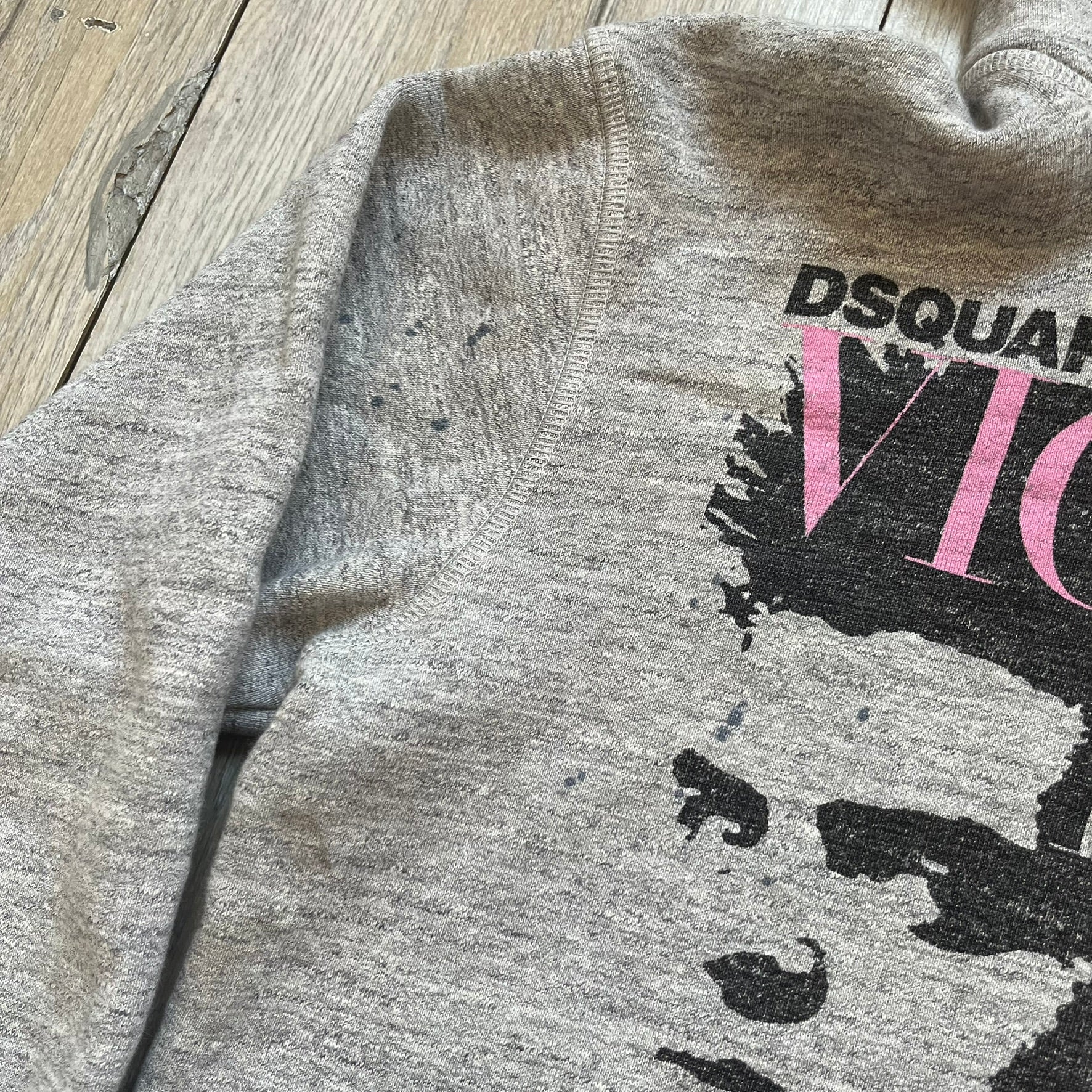 Dsquared2 Vicious Bros SS2019 Hoodie is a grey cotton hoodie made in Italy. It features a hood with drawstring fastening, long sleeves, and ribbed cuffs and hem. The front includes a printed graphic of two outlined figures with a small Dsquared2 logo and pink “Vicious Bros” text above them. The hoodie has intentional distressing throughout the fabric.
