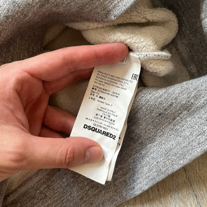 Dsquared2 Vicious Bros SS2019 Hoodie is a grey cotton hoodie made in Italy. It features a hood with drawstring fastening, long sleeves, and ribbed cuffs and hem. The front includes a printed graphic of two outlined figures with a small Dsquared2 logo and pink “Vicious Bros” text above them. The hoodie has intentional distressing throughout the fabric.
