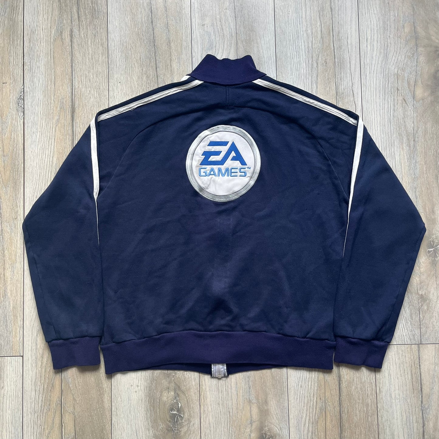 EA Sports 2000s Track Jacket is a navy track jacket with a full white zip closure, white two stripes running down both sleeves, and a fold-down collar. The back features a large stitched EA GAMES circular logo patch.&nbsp;