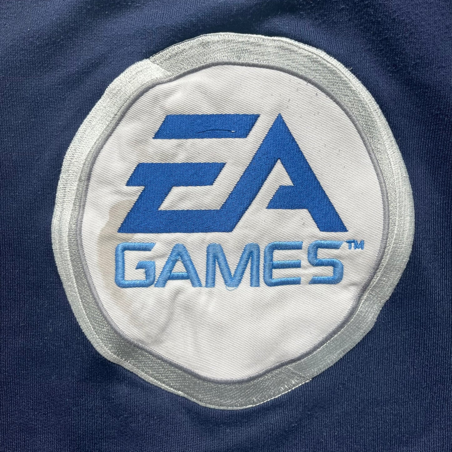 EA Sports 2000s Track Jacket is a navy track jacket with a full white zip closure, white two stripes running down both sleeves, and a fold-down collar. The back features a large stitched EA GAMES circular logo patch.&nbsp;