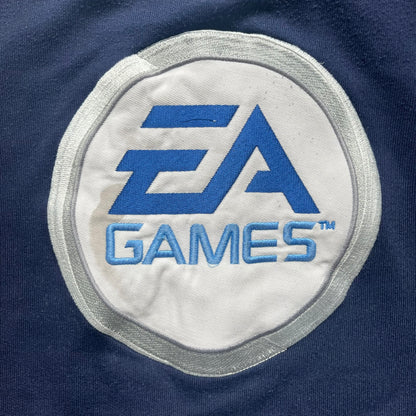 EA Sports 2000s Track Jacket is a navy track jacket with a full white zip closure, white two stripes running down both sleeves, and a fold-down collar. The back features a large stitched EA GAMES circular logo patch.&nbsp;