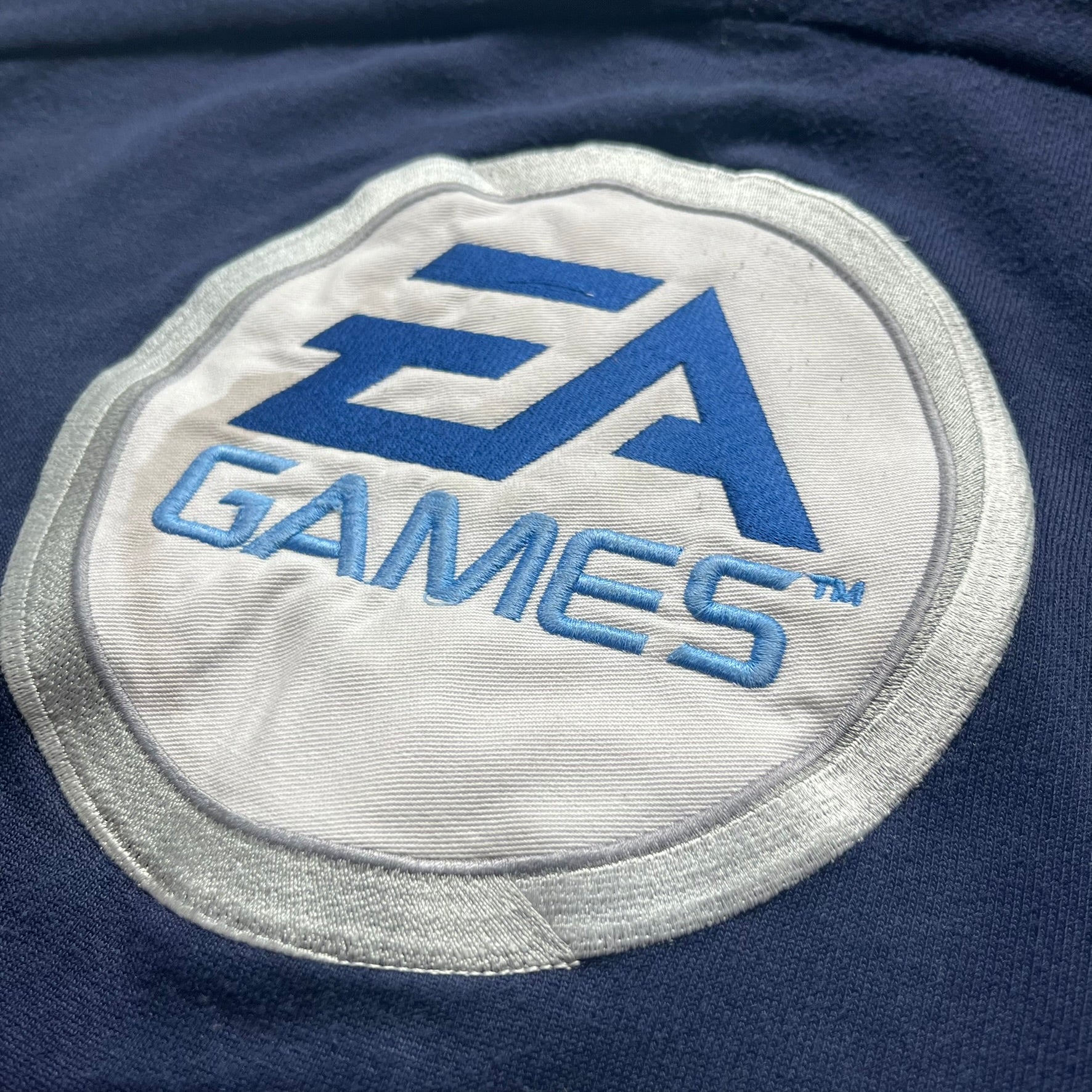 EA Sports 2000s Track Jacket is a navy track jacket with a full white zip closure, white two stripes running down both sleeves, and a fold-down collar. The back features a large stitched EA GAMES circular logo patch.&nbsp;