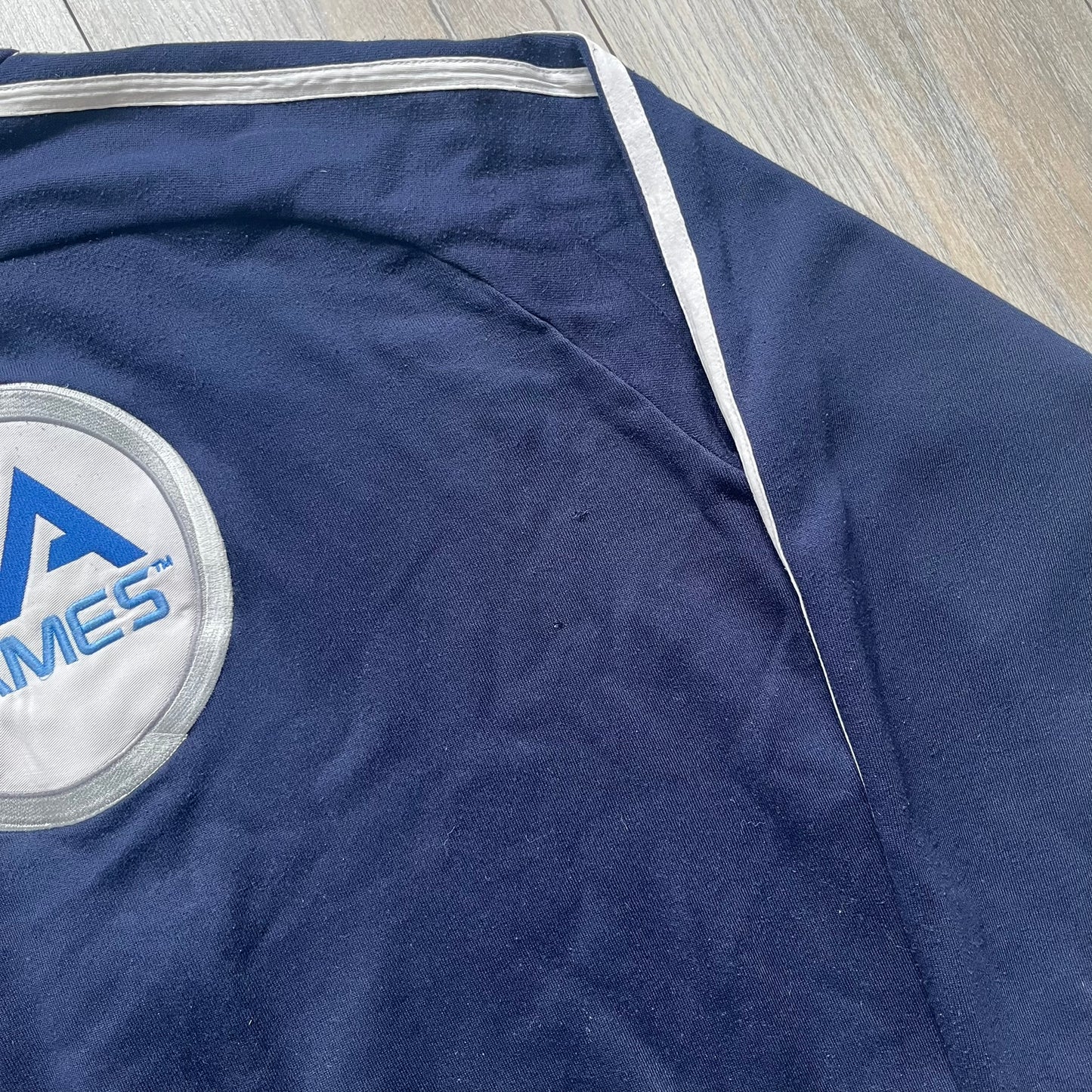 EA Sports 2000s Track Jacket is a navy track jacket with a full white zip closure, white two stripes running down both sleeves, and a fold-down collar. The back features a large stitched EA GAMES circular logo patch.&nbsp;