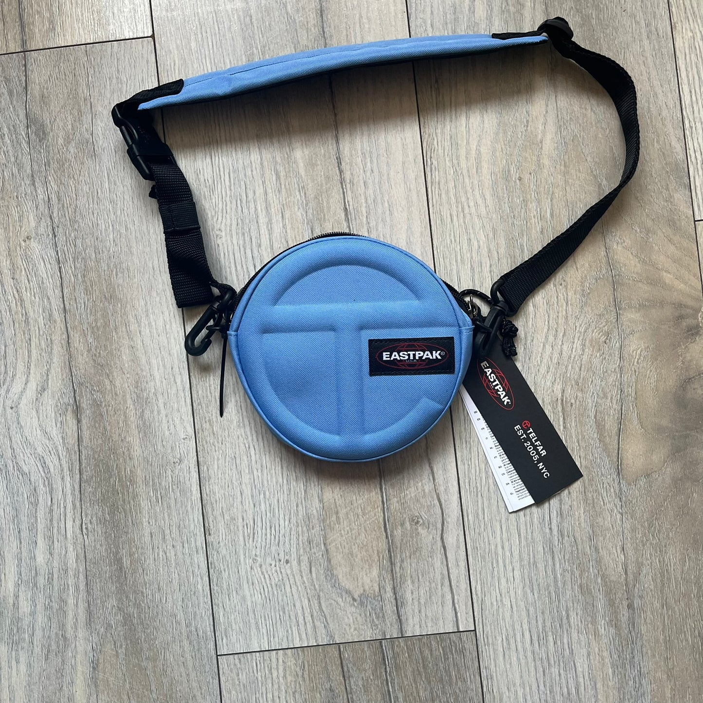 Eastpak x Telfar Circle Blue Shoulder Bag is crafted from polyester tactical canvas in a Cerulean colorway with black-tone hardware. The bag features woven hand straps, a removable and adjustable padded shoulder strap for crossbody or waist wear, and an embossed Telfar logo on the exterior. Released for SS24 on 06/14/2024 with a retail price of $110, the interior includes zip pockets and mesh compartments for organized storage.