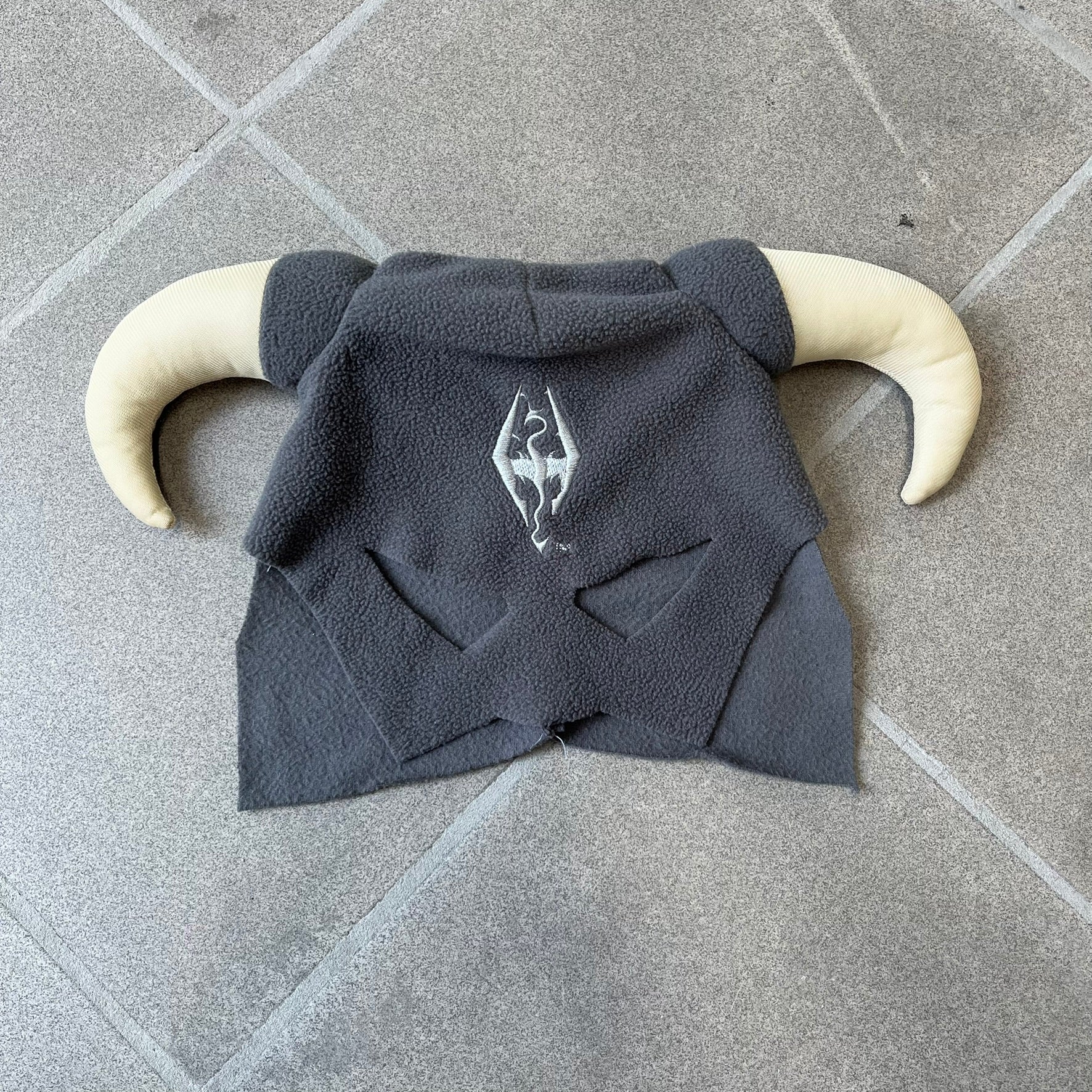 The Elder Scrolls V: Skyrim 2011 Dragonborn Helmet Hat is a grey fleece helmet-style beanie with large cream-colored curved horns and eye openings. The front features an embroidered light grey Skyrim dragon emblem on the forehead. Originally distributed by Bethesda as a promotional item at PAX Prime in 2011.
