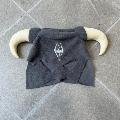 The Elder Scrolls V: Skyrim 2011 Dragonborn Helmet Hat is a grey fleece helmet-style beanie with large cream-colored curved horns and eye openings. The front features an embroidered light grey Skyrim dragon emblem on the forehead. Originally distributed by Bethesda as a promotional item at PAX Prime in 2011.
