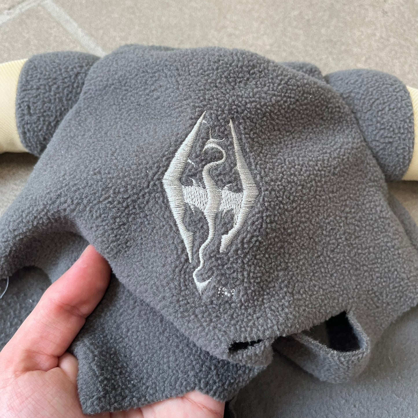 The Elder Scrolls V: Skyrim 2011 Dragonborn Helmet Hat is a grey fleece helmet-style beanie with large cream-colored curved horns and eye openings. The front features an embroidered light grey Skyrim dragon emblem on the forehead. Originally distributed by Bethesda as a promotional item at PAX Prime in 2011.
