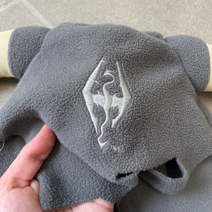 The Elder Scrolls V: Skyrim 2011 Dragonborn Helmet Hat is a grey fleece helmet-style beanie with large cream-colored curved horns and eye openings. The front features an embroidered light grey Skyrim dragon emblem on the forehead. Originally distributed by Bethesda as a promotional item at PAX Prime in 2011.
